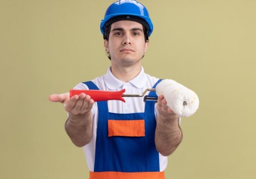 young-builder-man-construction-uniform-safety-helmet-demonstrating-paint-roller-looking-front-with-serious-face-standing-green-wall_141793-71906